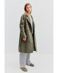 A person poses against a plain background, donned in an oversized fit that includes the chic Kule's The Rox trench coat made from water-resistant fabric in olive green. It's stylishly layered over a grey sweater and wide-leg pants, complemented by brown shoes to complete the look.