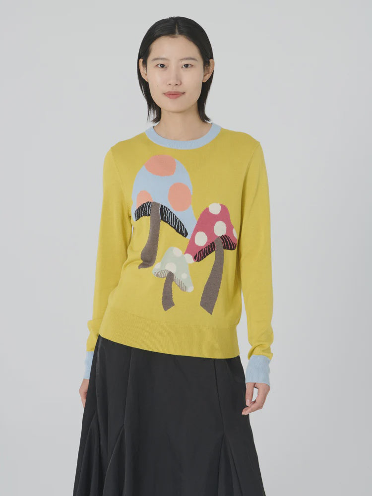 Dressed in Lingua Franca's "Mushroom Crew Lemon Drop," a whimsical sweater adorned with intarsia knit magic mushrooms, a person stands against a neutral background. The sweater features light blue cuffs and neckline, pairing perfectly with a black skirt for an ensemble that is both stylish and enchanting.