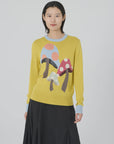 Dressed in Lingua Franca's "Mushroom Crew Lemon Drop," a whimsical sweater adorned with intarsia knit magic mushrooms, a person stands against a neutral background. The sweater features light blue cuffs and neckline, pairing perfectly with a black skirt for an ensemble that is both stylish and enchanting.