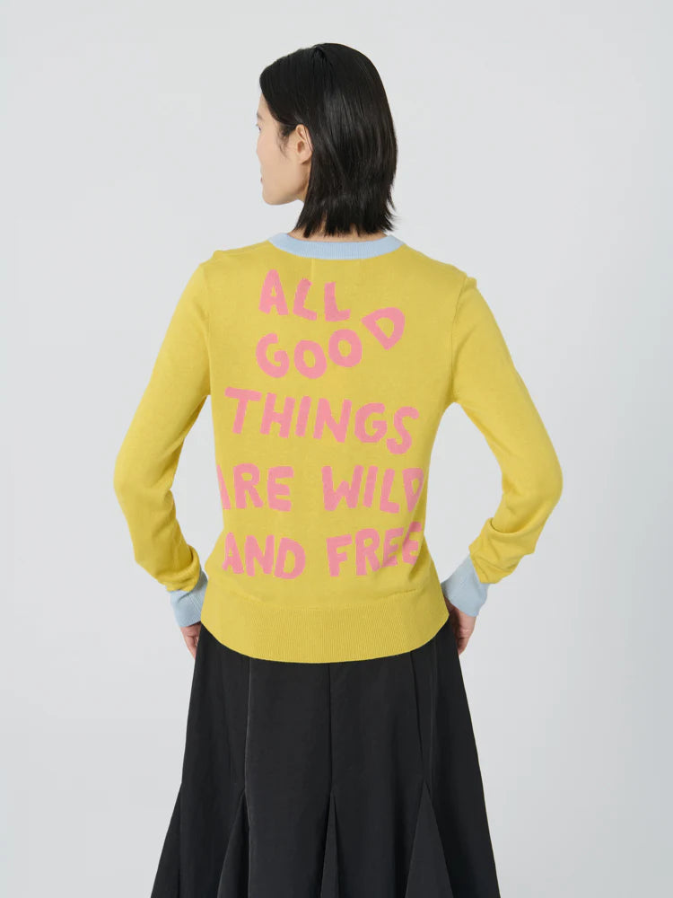 A person with short dark hair and facing away from the camera is wearing the Lingua Franca "Mushroom Crew Lemon Drop" sweater in yellow, which features pink intarsia knit text: "ALL GOOD THINGS ARE WILD AND FREE." They are paired with a black skirt and standing against a plain light gray background.