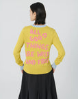 A person with short dark hair and facing away from the camera is wearing the Lingua Franca "Mushroom Crew Lemon Drop" sweater in yellow, which features pink intarsia knit text: "ALL GOOD THINGS ARE WILD AND FREE." They are paired with a black skirt and standing against a plain light gray background.