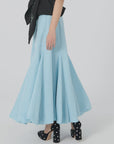 A person wearing the Donna Bubble Skirt by Lingua Franca in a flowing light blue hue is captured mid-step. They pair it with a black sleeveless top featuring a large bow, and walk confidently in black high heels embellished with white studs against a plain white backdrop.