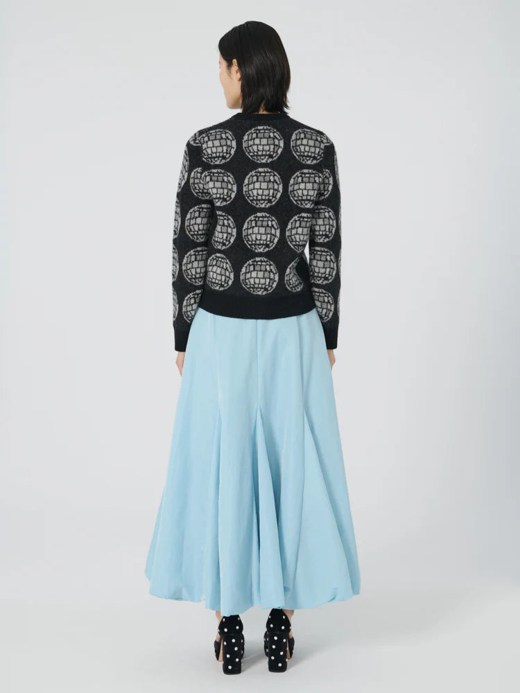 A person with shoulder-length dark hair is adorned in a black sweater showcasing an embroidered phrase, paired elegantly with the Lingua Franca's Donna Bubble Skirt in a long, flowing light blue hue. They face away from the camera against a simple white background, effortlessly combining style and understated elegance.