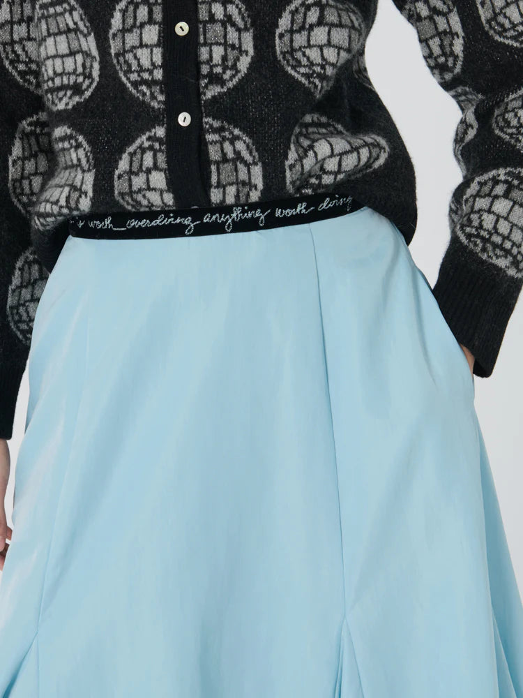 The person is dressed in a black sweater adorned with a globe design and a light blue Lingua Franca Donna Bubble Skirt, which showcases a velvet waistband with the phrase "work, coaching, anything worth doing.