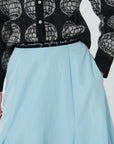 The person is dressed in a black sweater adorned with a globe design and a light blue Lingua Franca Donna Bubble Skirt, which showcases a velvet waistband with the phrase "work, coaching, anything worth doing.