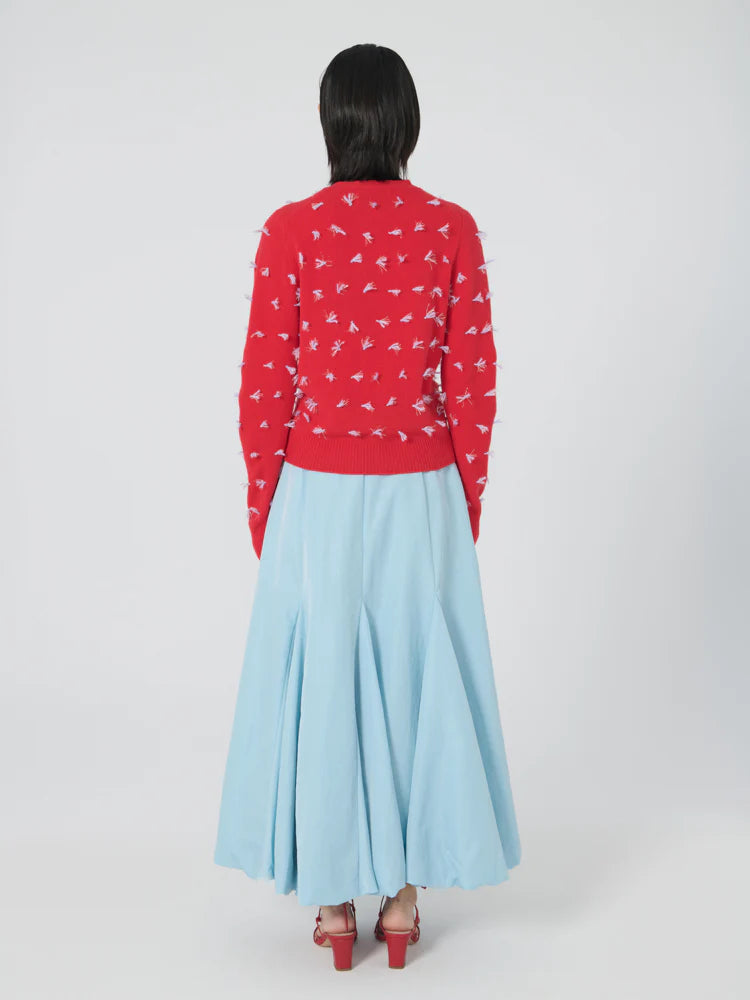 A person with shoulder-length dark hair is wearing a red Lingua Franca Diana Cardigan adorned with small white prints and paired with a long, light blue skirt crafted from soft merino wool. They are positioned against a plain white background, facing away from the camera. The ensemble is completed with red shoes.