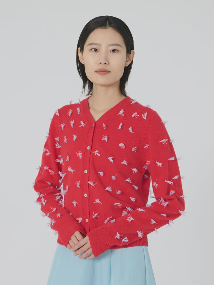 A person with short dark hair is wearing the Diana Cardigan in red by Lingua Franca, featuring white and purple embroidered details, paired with a light blue skirt. The background is plain white.