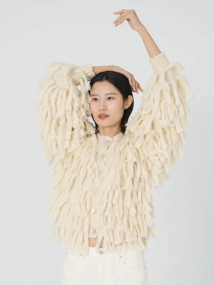 A person stands against a plain background, wearing the Cher Fringe Jacket by Lingua Franca, with its chunky cream-colored alpaca blend and textured, fluffy details. They strike a relaxed pose with one arm raised above their head while the other is bent and resting on their head.