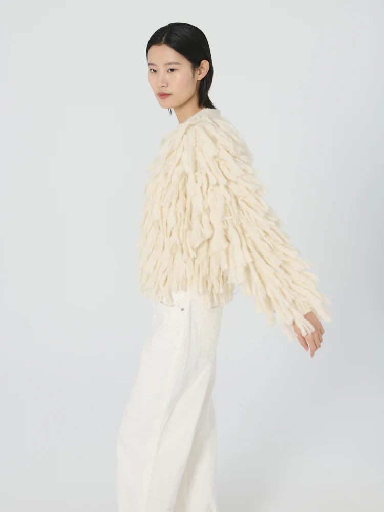 A person with short black hair is wearing a cream-colored alpaca blend sweater, featuring long fringed accents inspired by the Cher Fringe Jacket from Lingua Franca, paired with white pants. They are standing against a plain light backdrop, slightly turned to the side.