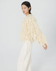 A person with short black hair is wearing a cream-colored alpaca blend sweater, featuring long fringed accents inspired by the Cher Fringe Jacket from Lingua Franca, paired with white pants. They are standing against a plain light backdrop, slightly turned to the side.