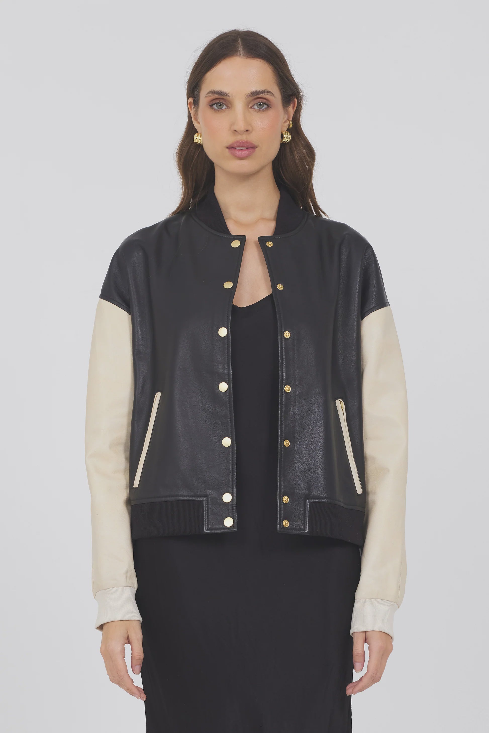 A woman with long brown hair faces forward, wearing the JAKETT Mae Washed Leather varsity jacket—black body, cream sleeves, gold buttons—styled over a black dress against a plain light background.