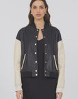A woman with long brown hair faces forward, wearing the JAKETT Mae Washed Leather varsity jacket—black body, cream sleeves, gold buttons—styled over a black dress against a plain light background.