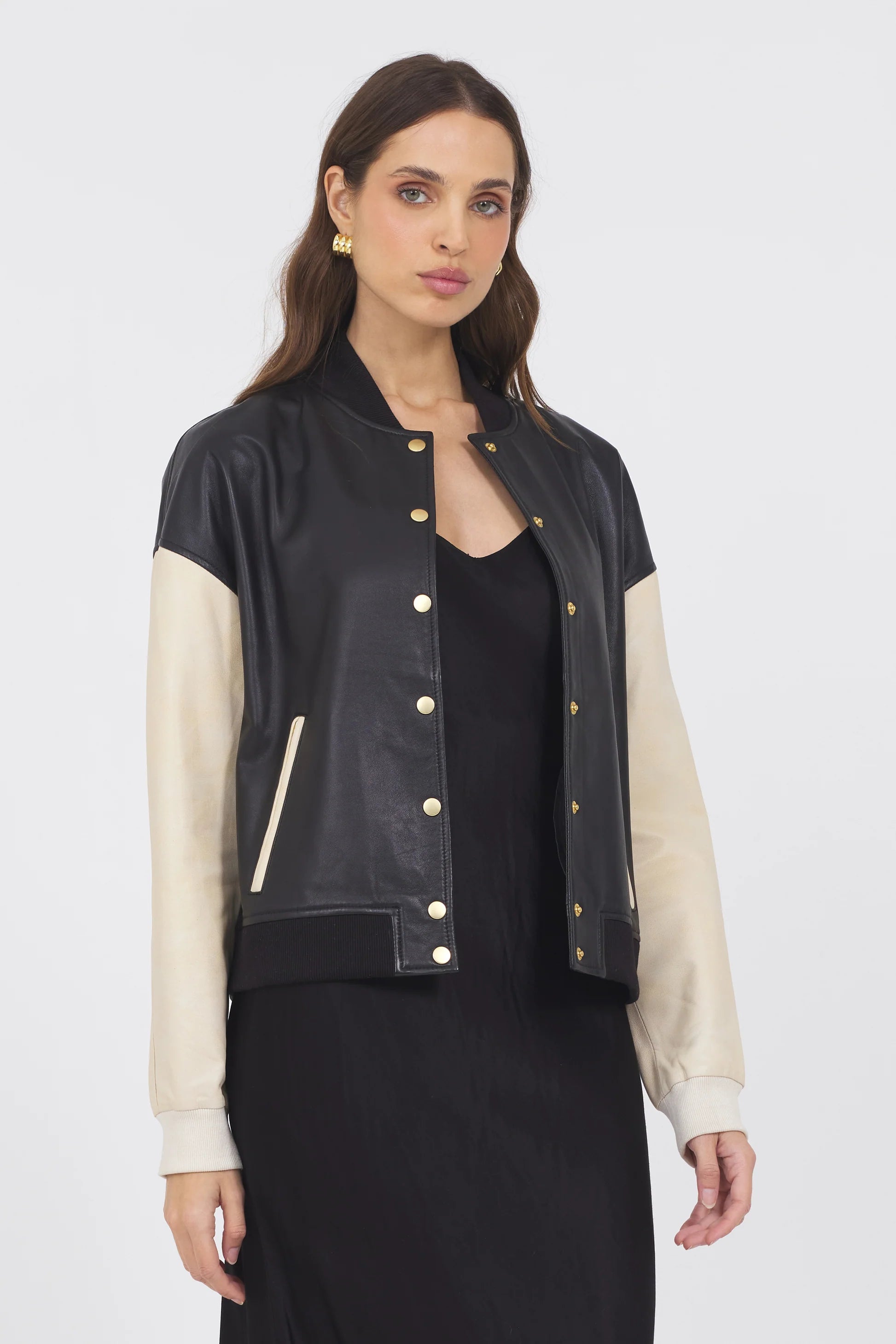 A woman with long brown hair wears a black dress and the JAKETT Mae Washed Leather jacket, featuring cream-colored sleeves and gold buttons, standing against a plain white background for a chic vintage look.