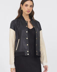 A woman with long brown hair wears a black dress and the JAKETT Mae Washed Leather jacket, featuring cream-colored sleeves and gold buttons, standing against a plain white background for a chic vintage look.