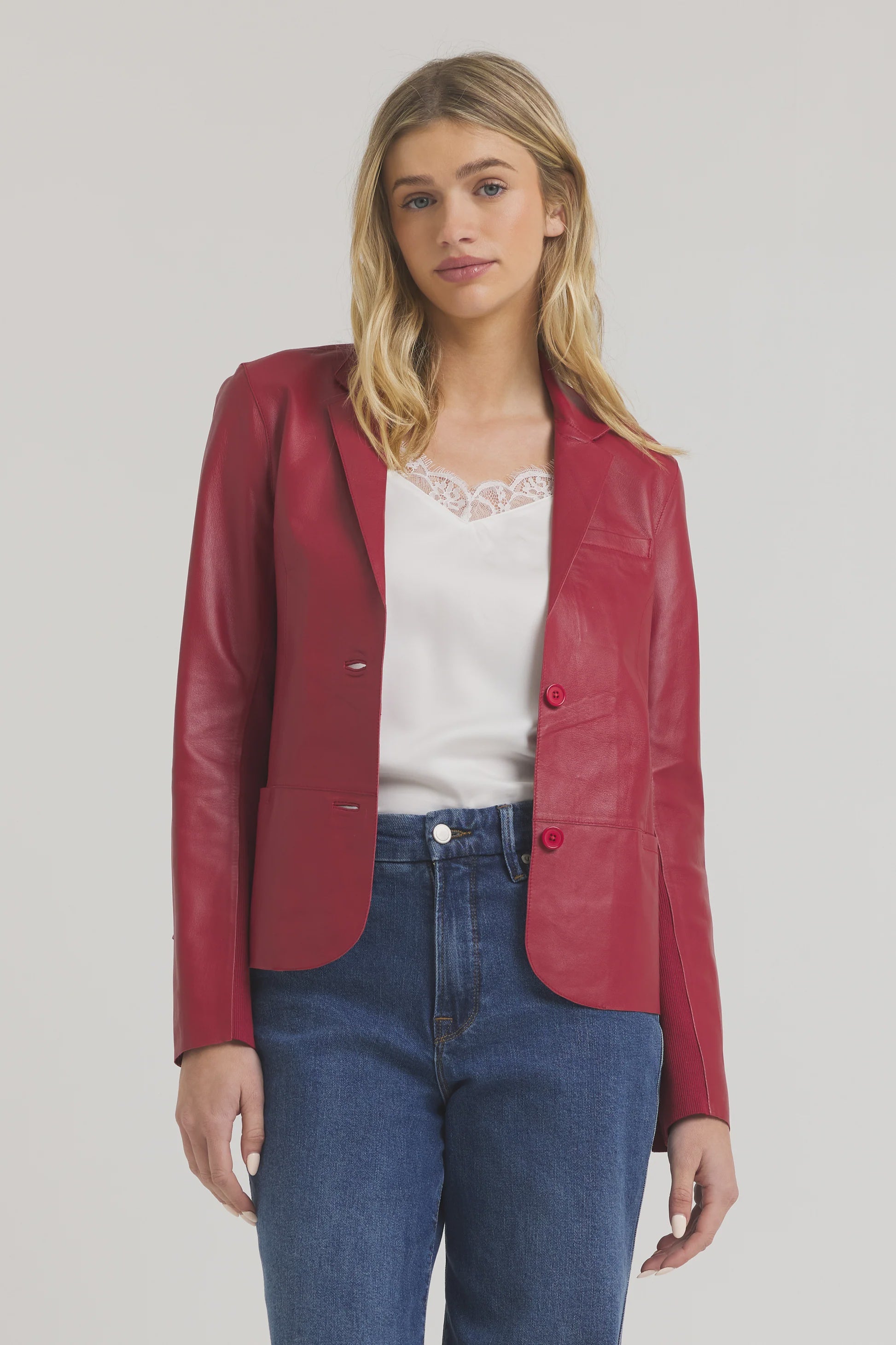 A woman wears the JAKETT Allison Burnished Leather red blazer over a white lace-trimmed top and blue jeans, standing against a plain light background with a neutral expression.