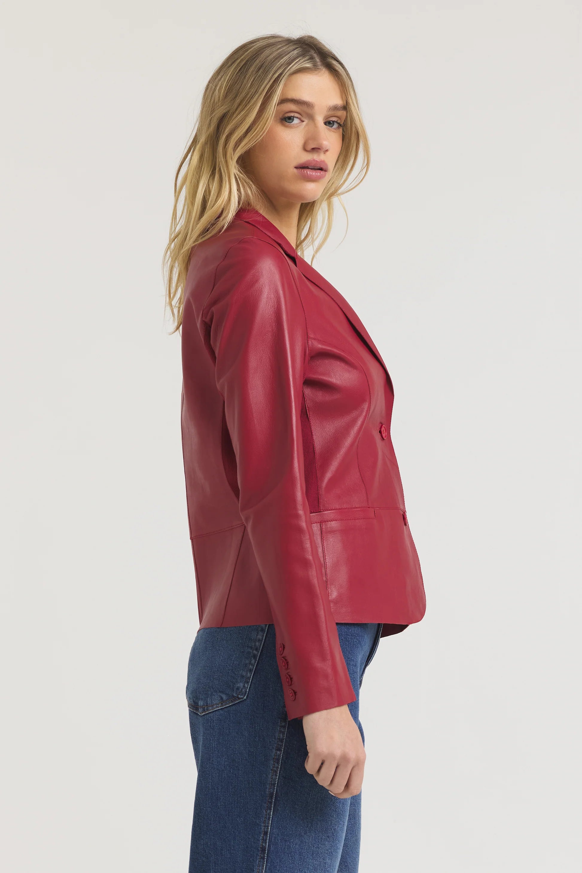 A person with long blonde hair, wearing JAKETT's Allison Burnished Leather red blazer and blue jeans, stands sideways against a plain light background.