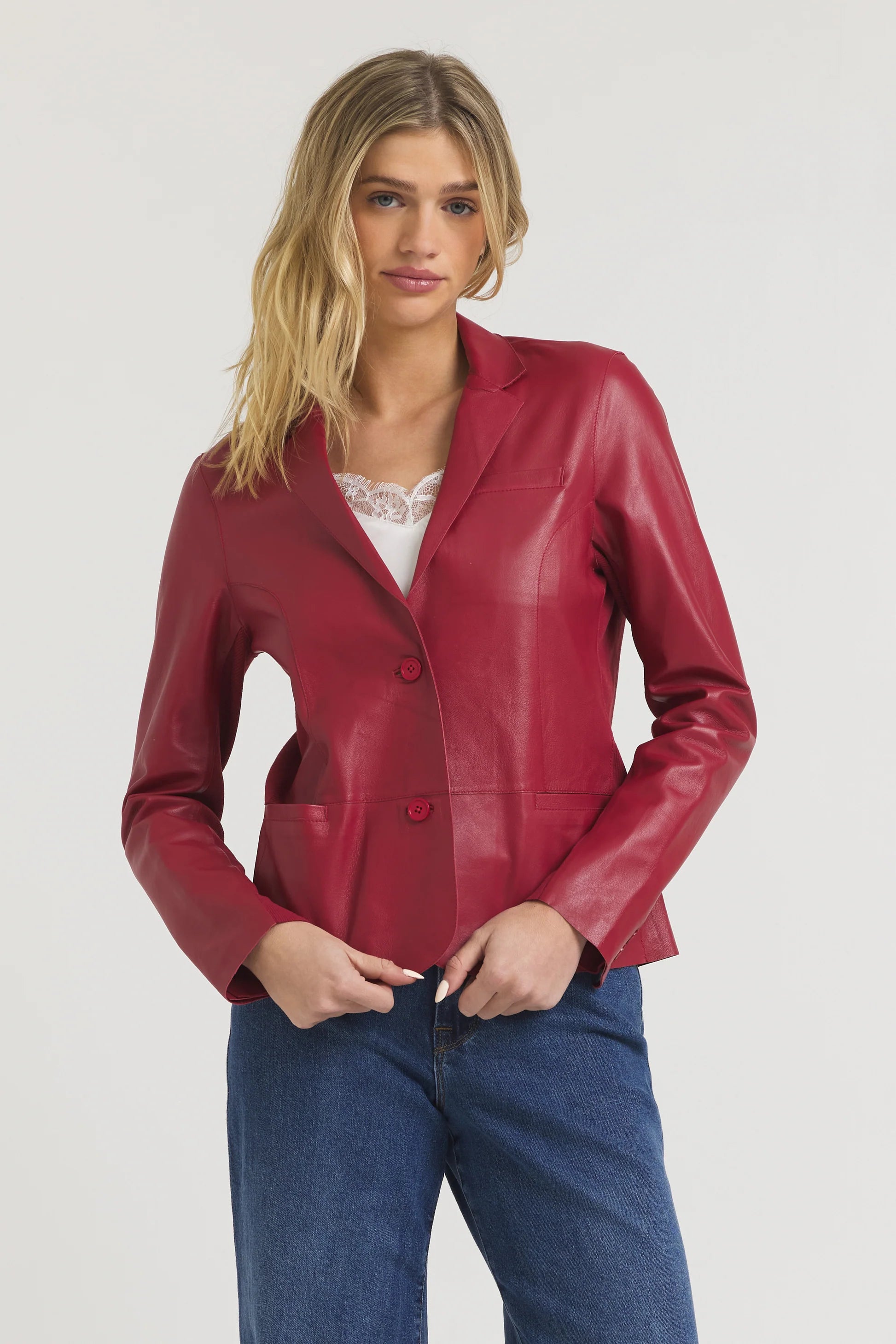 A woman stands against a plain light background, wearing the JAKETT Allison Burnished Leather red blazer over a white lace-trimmed top and blue jeans, her hands resting on the sides of the blazer.