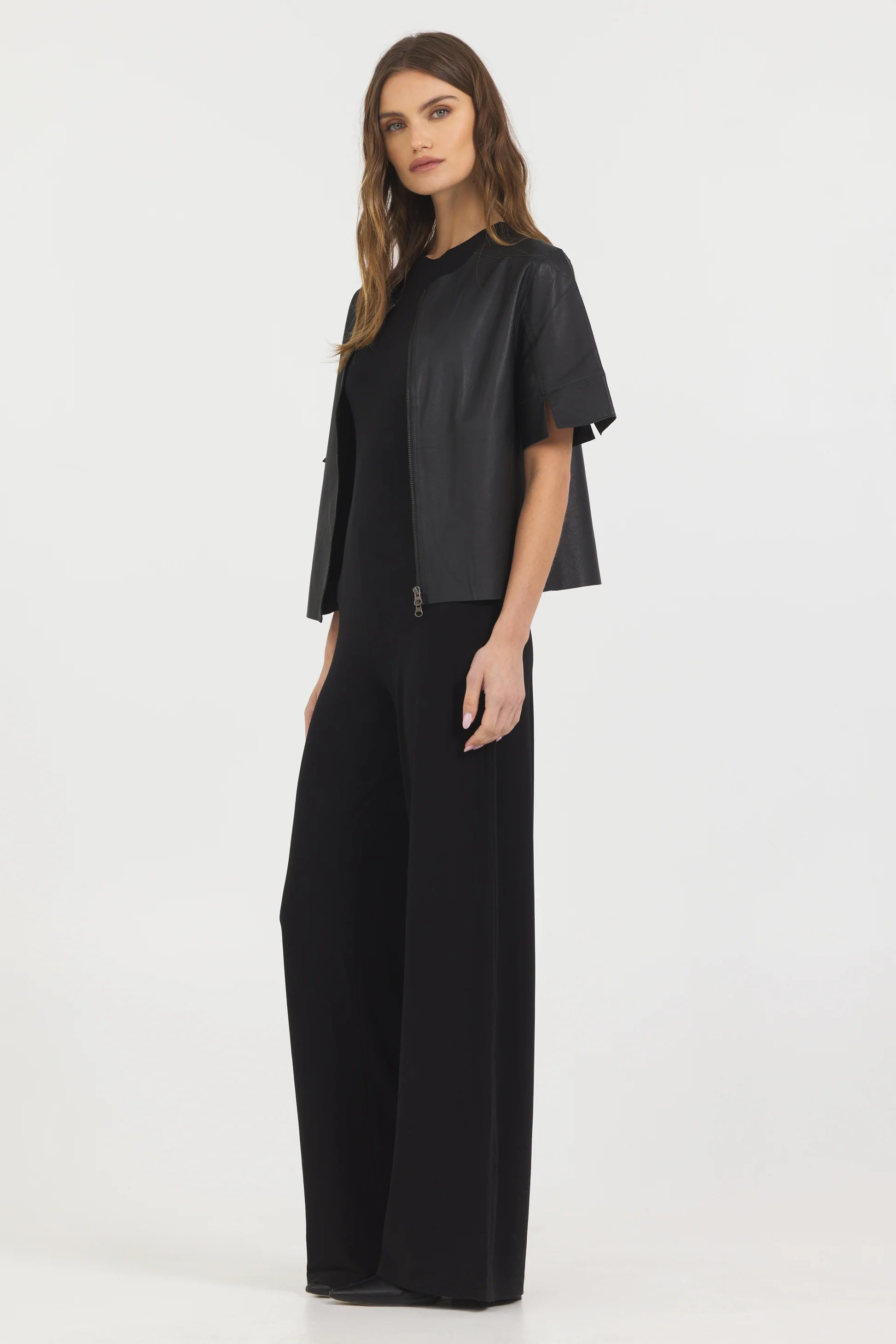 A woman with long wavy hair and a neutral expression wears the JAKETT Leather T-shirt over a black top and wide-leg black pants, standing against a plain white background.