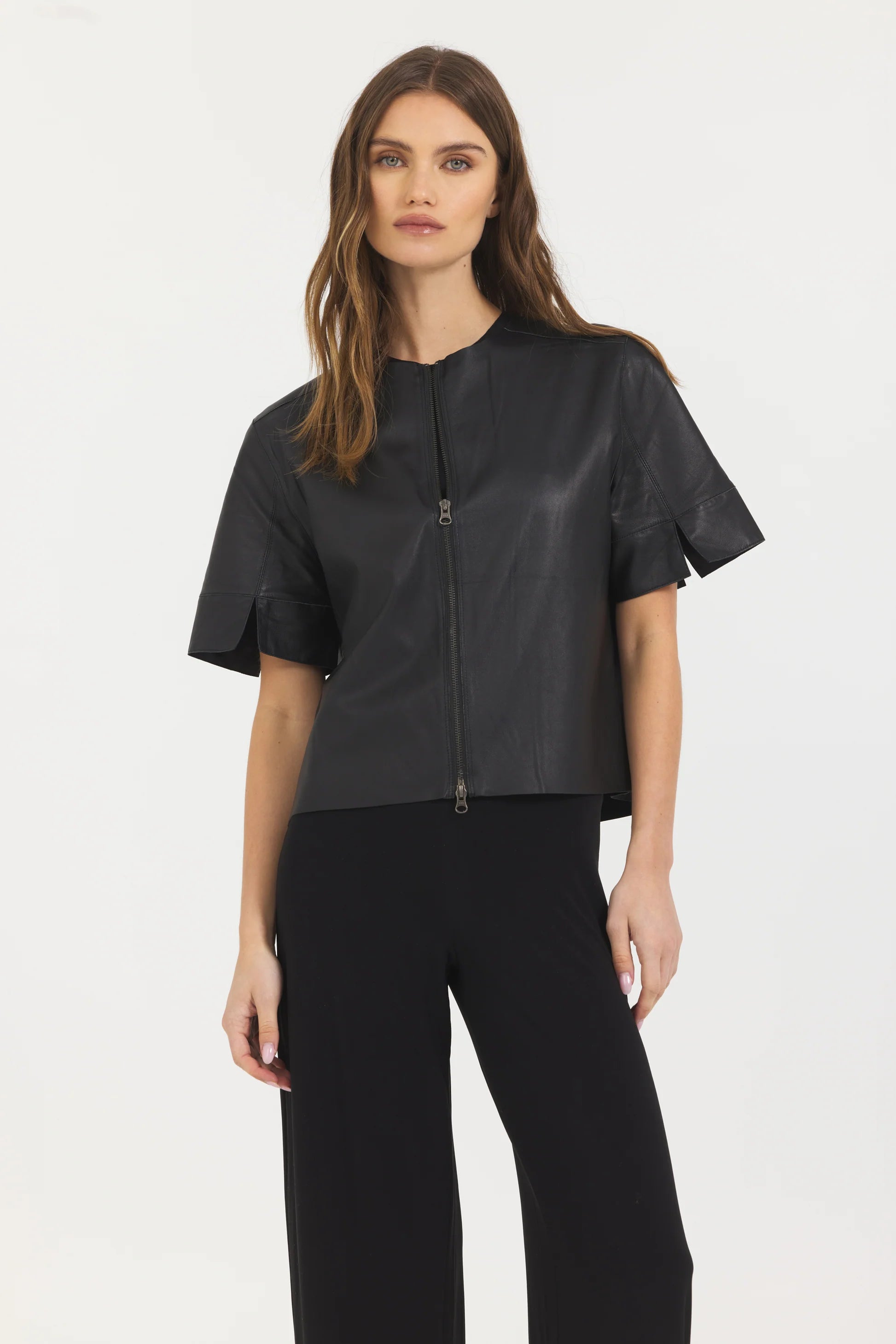 A woman with long brown hair models the JAKETT Leather T-shirt and black pants, standing against a plain white background.