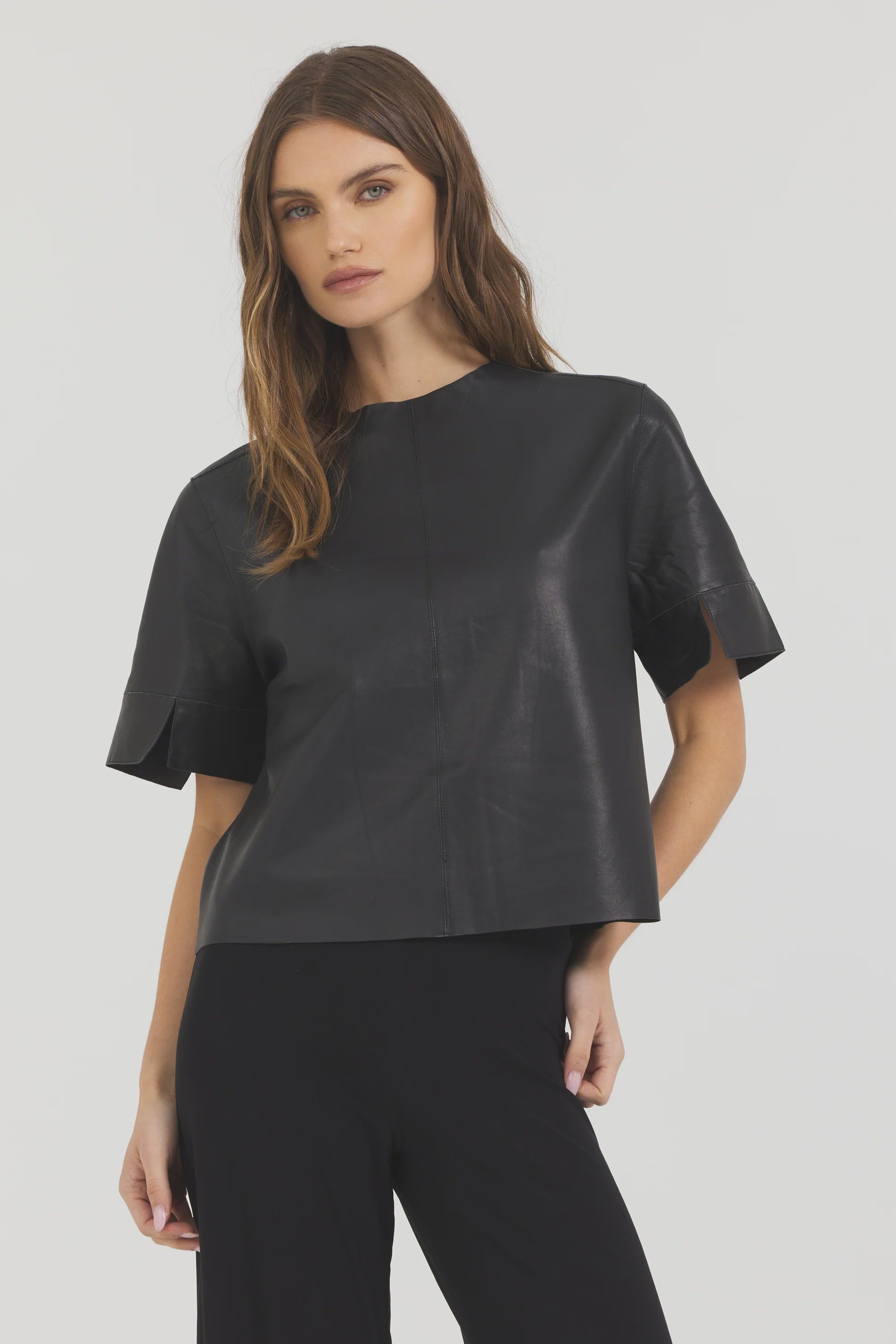 A woman with long, wavy brown hair poses against a plain background wearing the JAKETT Leather T-shirt and black pants, one hand resting on her hip.