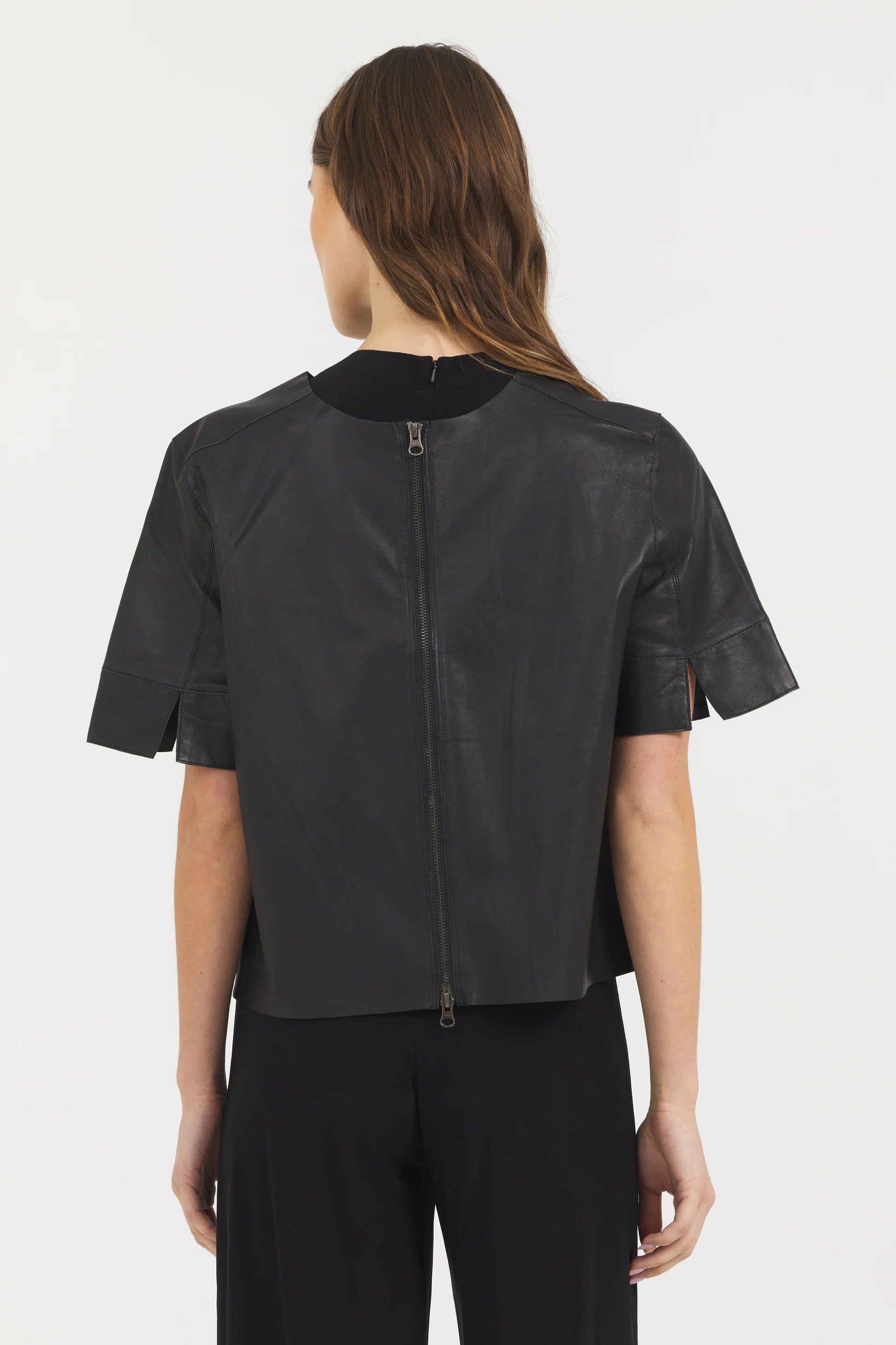 A woman with long brown hair is seen from behind wearing the JAKETT Leather T-shirt—an oversized, boxy black leather top with a center-back zipper—paired with black pants, set against a plain white background.