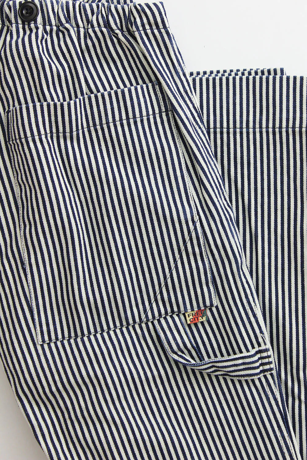 A close-up of the Indigo Stripe Sailor Pant by Free City (sparrow, LLC), featuring unisex blue and white vertical stripes, a front pocket with black button, and a small red and white "FRY ONE" tag on the seam.