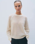 A person with light skin and curly hair pulled back wears the White + Warren Cashmere Mesh Sweatshirt in cream and black pants, standing against a plain light background with hands in pockets, looking at the camera.