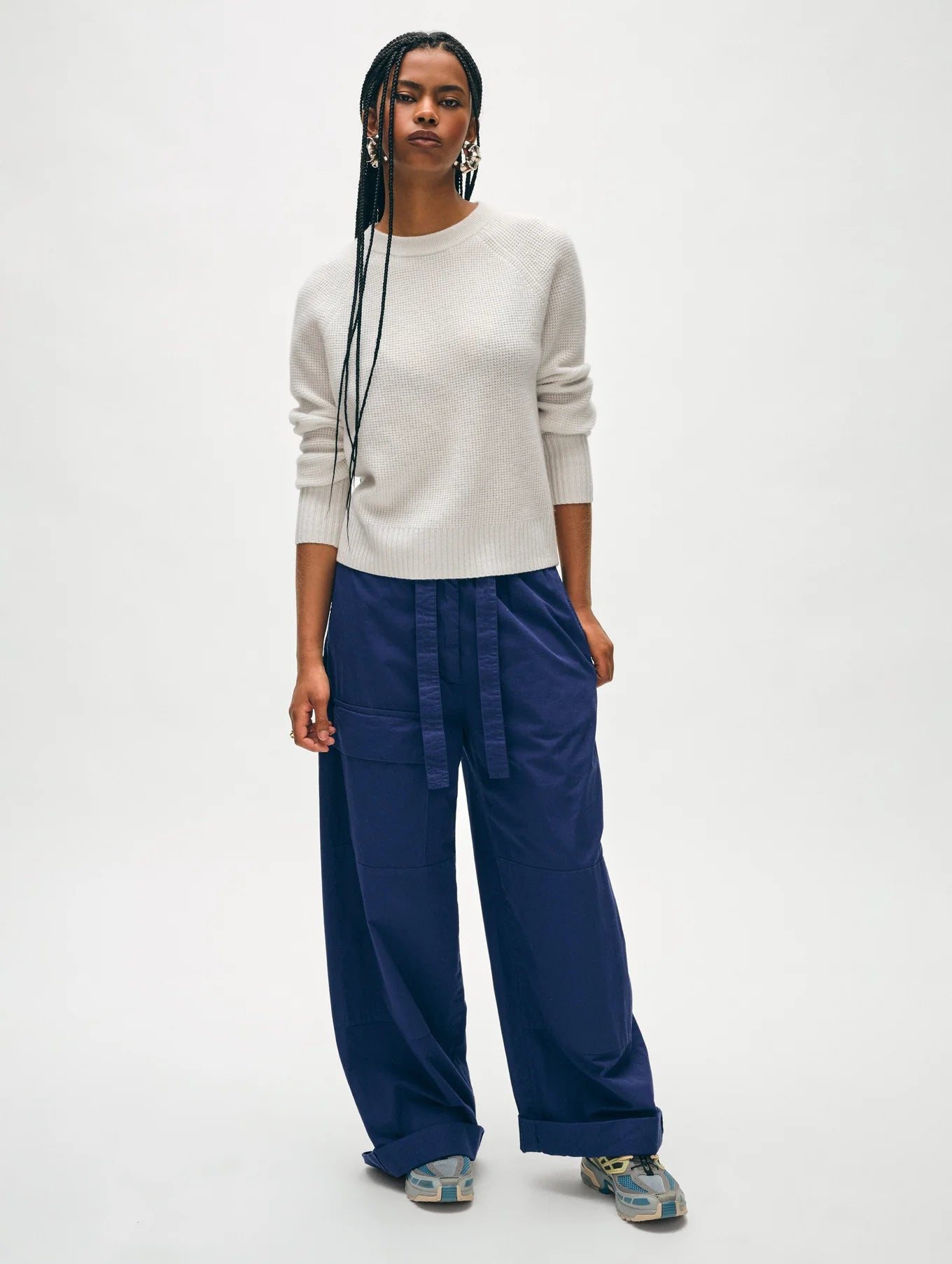 A person with long braided hair stands against a plain background, wearing the White + Warren Cashmere Waffle Sweatshirt in light gray, distinguished by its signature waffle-knit stitch. They pair it with wide-leg navy pants and sneakers, their hands resting relaxed at their sides and a neutral expression on their face.