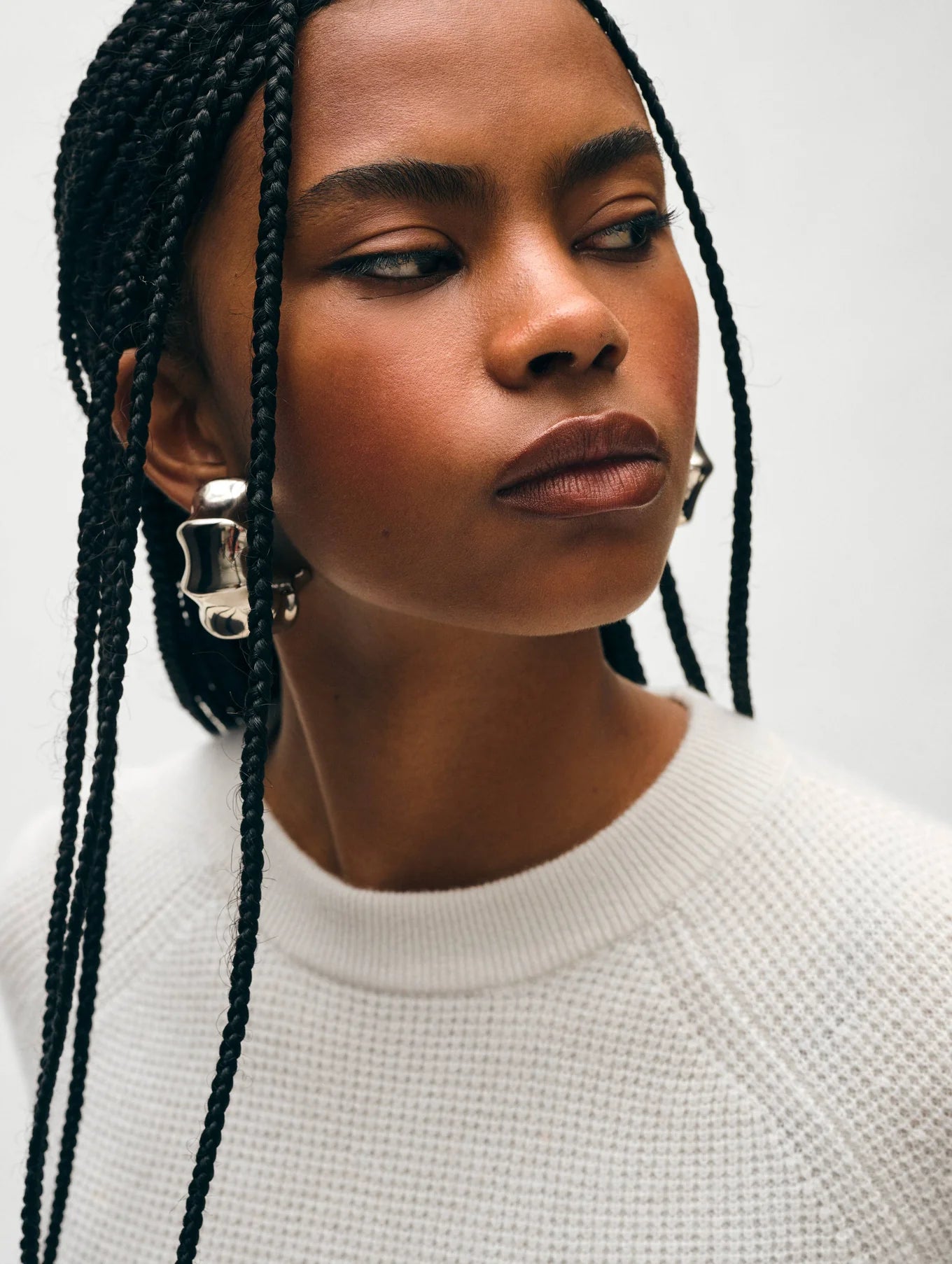 A person with long braided hair and large, metallic earrings glances to the side while wearing a Cashmere Waffle Sweatshirt from White + Warren. The textured white garment's relaxed fit enhances their style against a plain, neutral background.