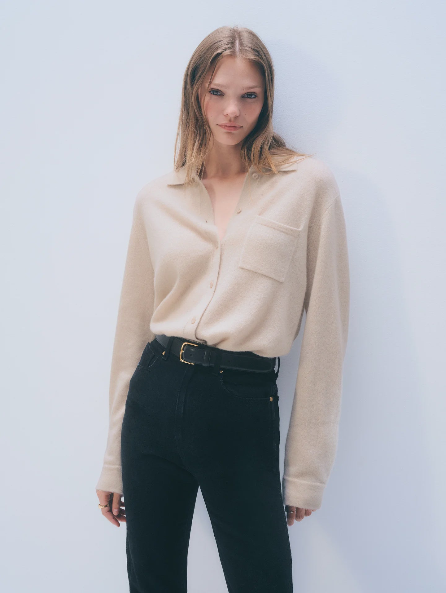 A woman with straight, light brown hair wears the White + Warren Cashmere Button Down in beige with a chest pocket and black high-waisted pants, standing against a plain light background.