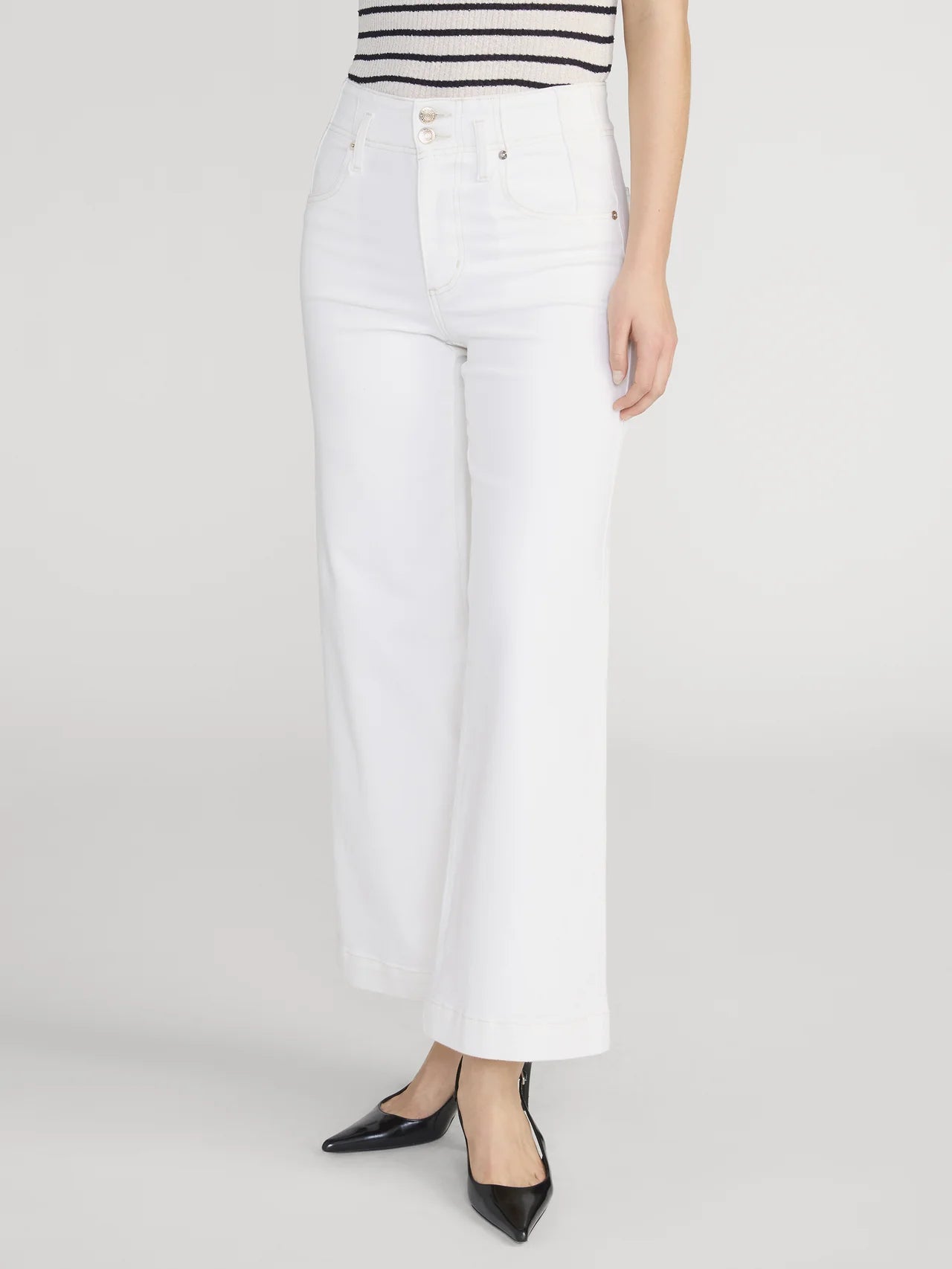 A person models FRAME’s Wide Leg Trouser Crop, featuring high-rise, wide-leg white jeans in super-stretch denim. Styled with a sleeveless striped top and black pointed-toe heels. Only the lower body is visible against a plain light backdrop.