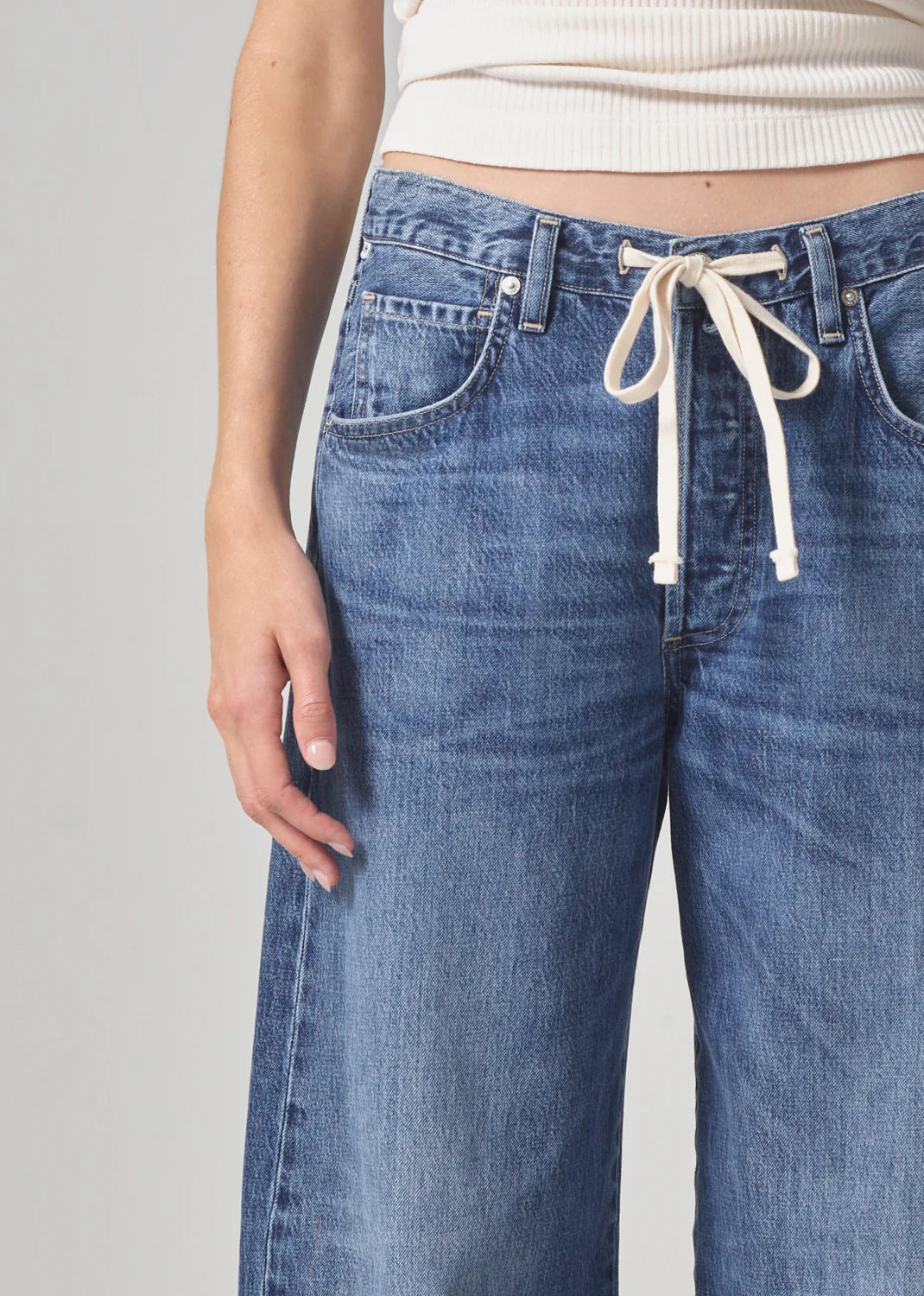 Close-up of someone wearing the Brynn Trouser in Atlantis from Citizens Of Humanity/AGOLDE, featuring wide-leg blue denim and a white drawstring waist, paired with a cream ribbed sweater. The person's left arm rests against a plain light gray background.