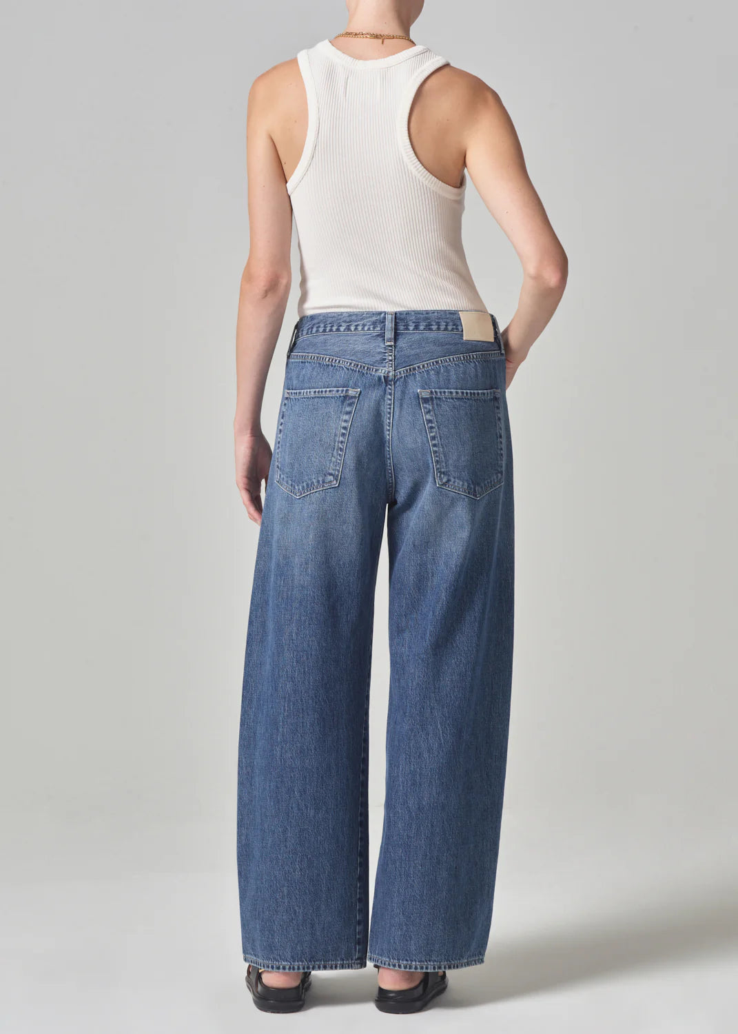 A person stands facing away, showcasing a white ribbed tank top paired with the brynn trouser in atlantis by Citizens Of Humanity/AGOLDE. The wide-leg blue jeans feature a stylish drawstring waist against a plain light gray background.