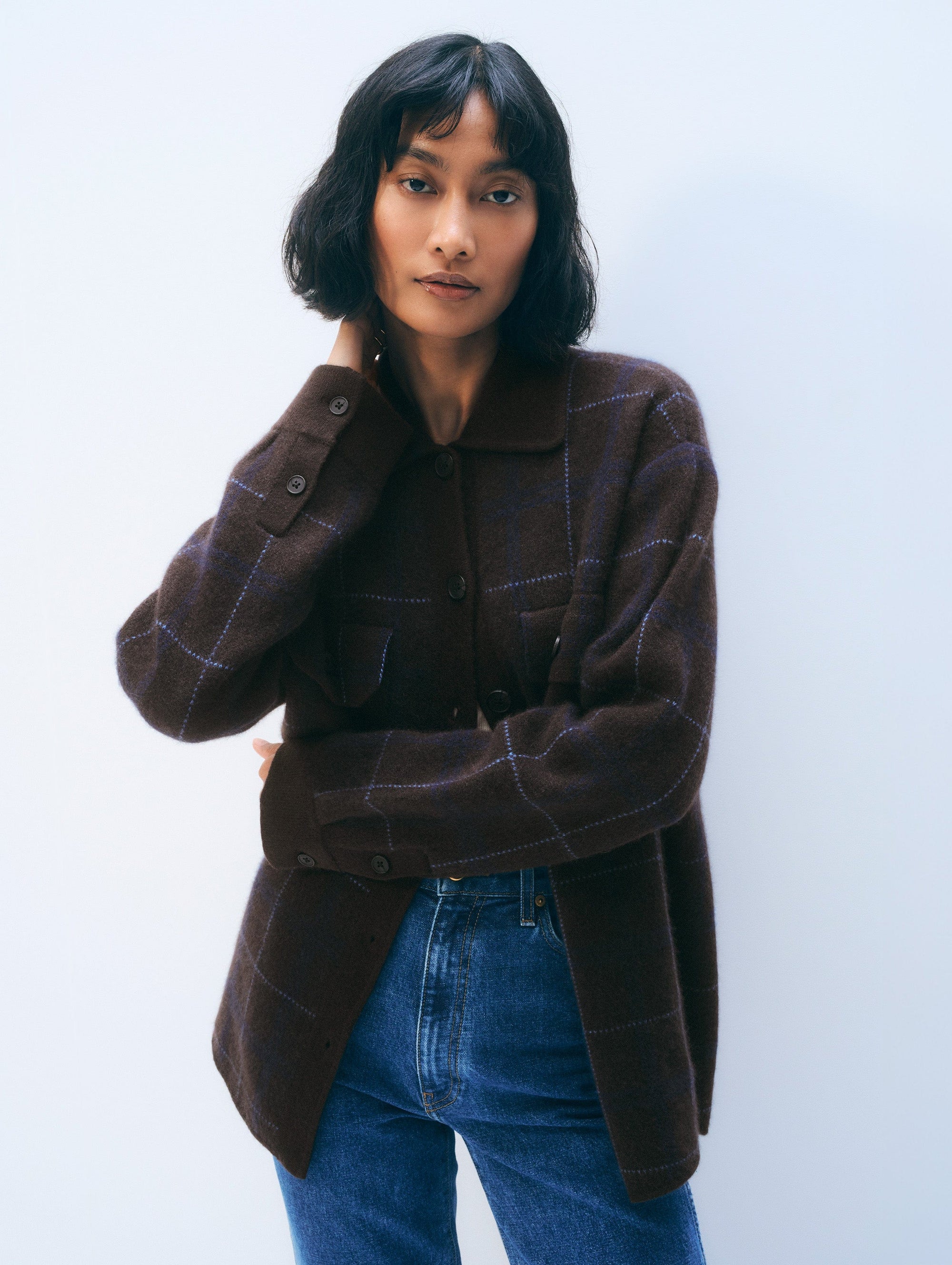 A woman with short black hair wears the White + Warren Cashmere Plaid Shacket with blue jeans, standing against a plain light background. She touches her neck and looks confidently at the camera.