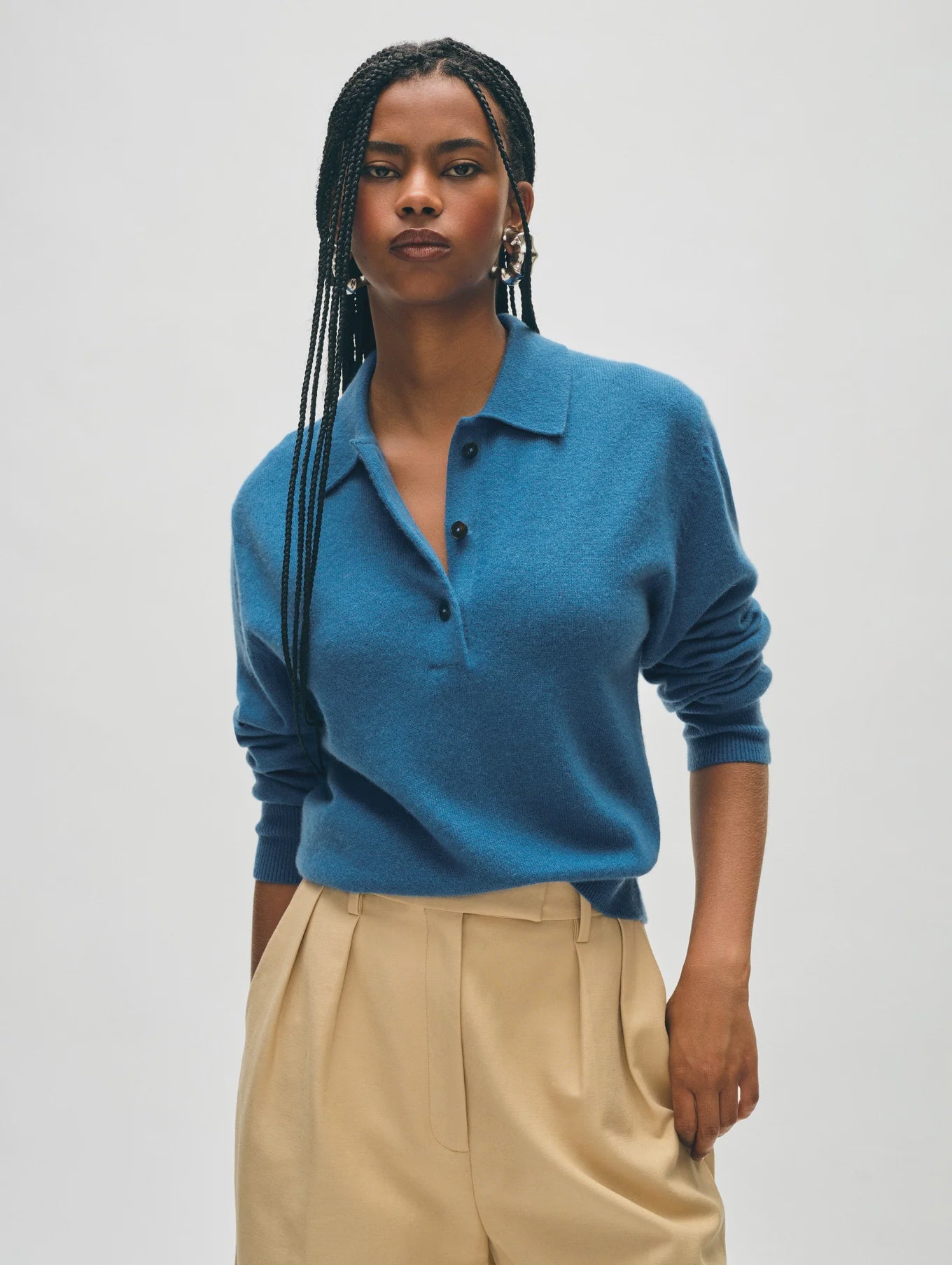 A person with long braided hair stands against a neutral background, wearing the Cashmere Button Polo from White + Warren and beige pleated pants. They have their hands at their sides and are looking confidently at the camera, showcasing a relaxed fit.