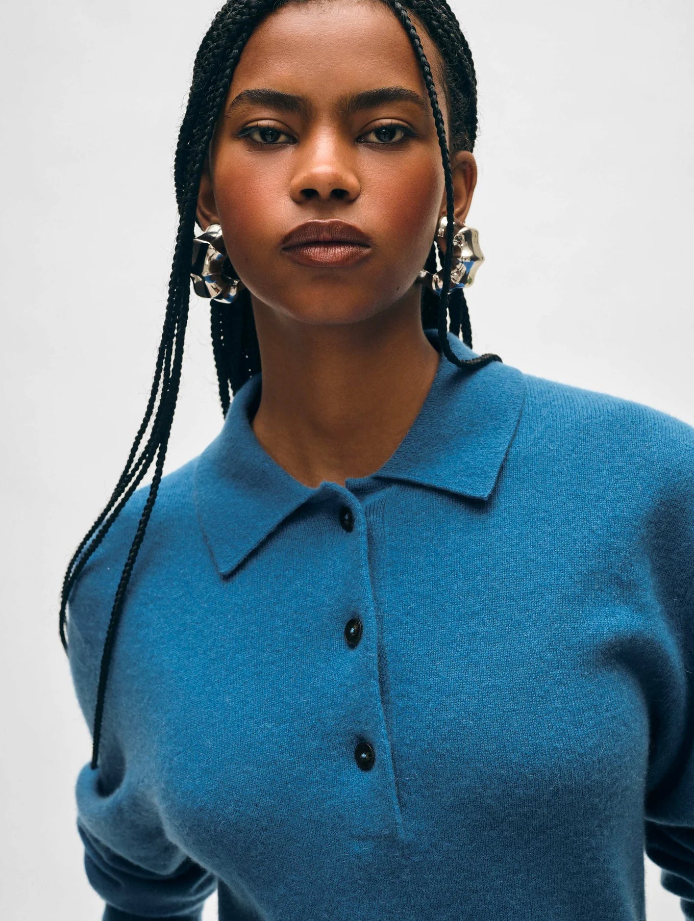 A person with long braided hair wears a Blue Cashmere Button Polo by White + Warren, adorned with ribbed trims, paired with large silver earrings. They stand against a plain background, looking directly at the camera with a confident expression.