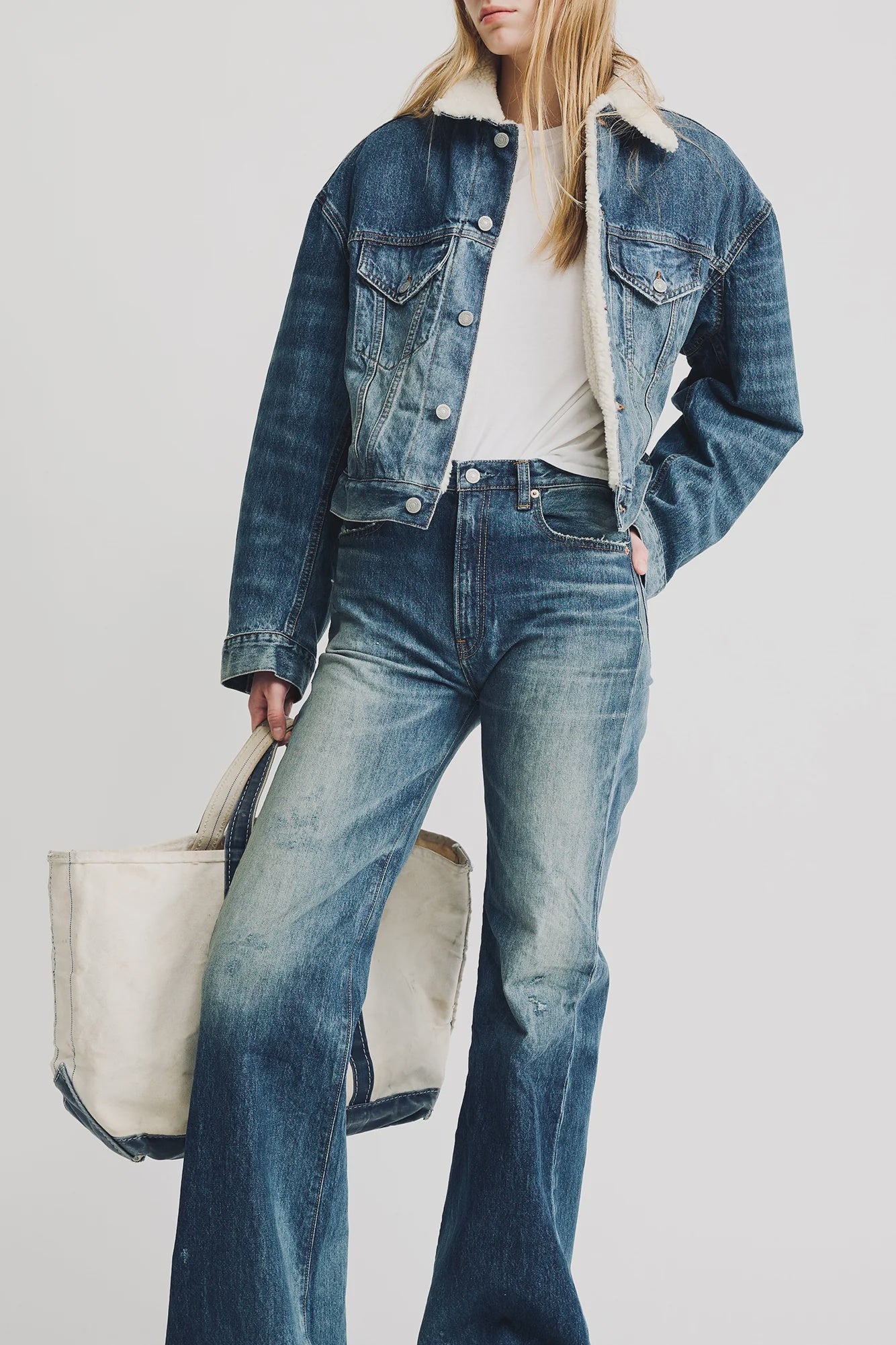 A woman wears the Denimist IRIS BOXY TRUCKER JACKET in indigo denim with sherpa lining, paired with matching high-waisted flared jeans and a white tee. She carries a large off-white tote; her hair is loose and her face partly out of frame.