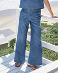 Wearing Frank & Eileen's Madeline Patch Pocket Pant in indigo denim and flip-flops, a person stands on a white wooden balcony overlooking the beach and surrounding greenery.