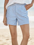A person wears Frank & Eileen’s Waterford Short in blue and white gingham with a wide-leg fit and a white top, standing on a sandy beach with the ocean blurred in the background. Only the lower torso and upper legs are shown.