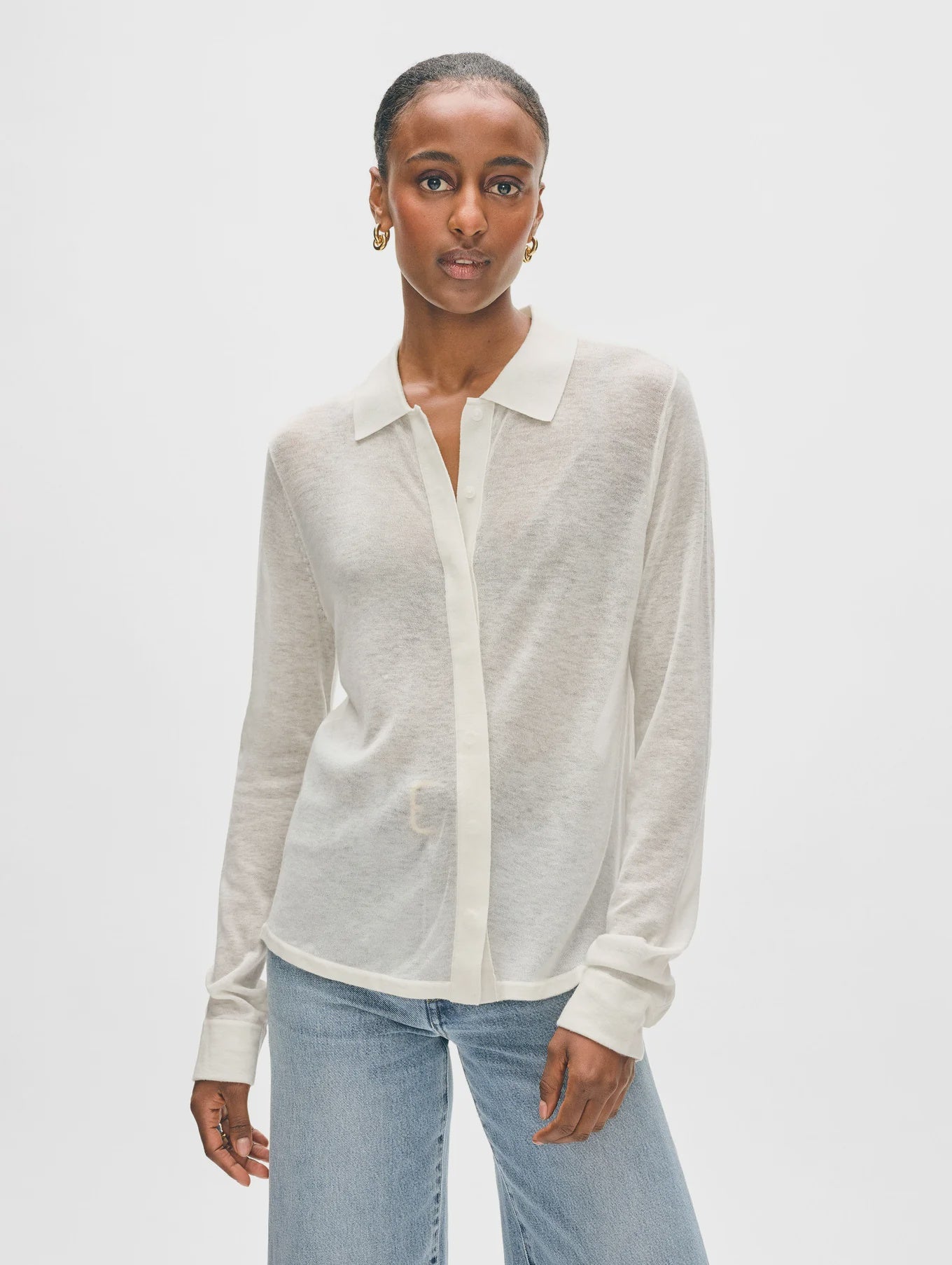 A person in a superfine cashmere button-down by White + Warren and blue jeans stands against a white background. They have short hair and wear hoop earrings.