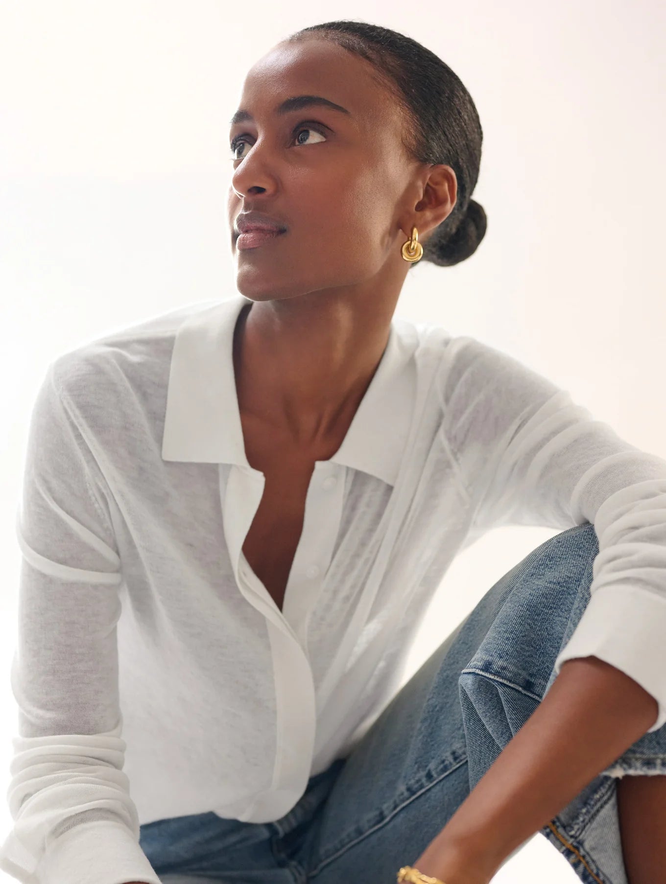 A person with hair pulled back sits looking sideways, wearing a superfine cashmere button-down from White + Warren and light blue jeans. Gold hoop earrings and a gold bracelet add elegance, enhanced by soft lighting for a refined ambiance.