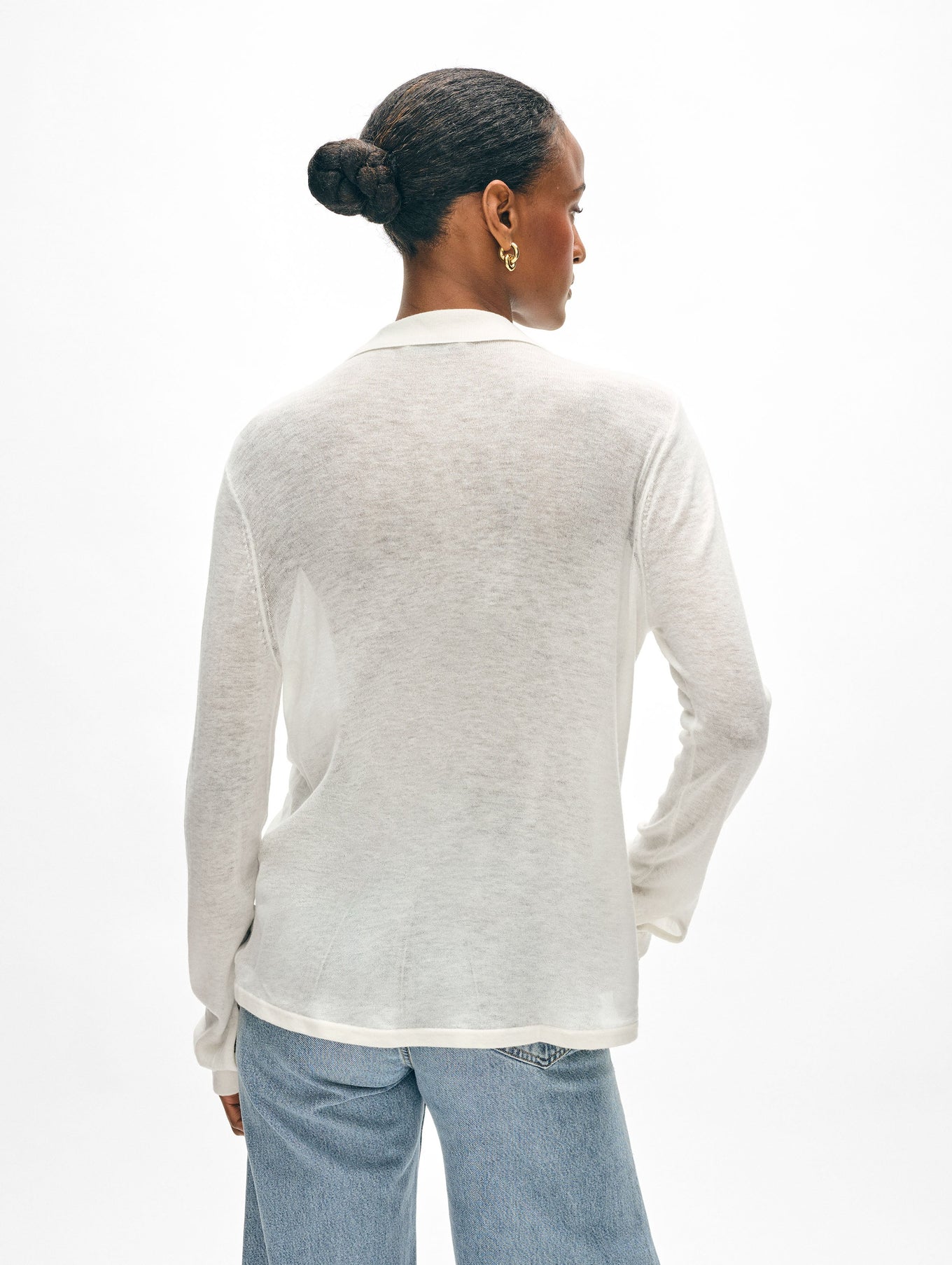 A person with hair in a bun and hoop earrings stands with their back to the camera, wearing a sheer superfine cashmere button down by White + Warren, paired with light blue jeans. The plain white background subtly highlights the elegance of their ensemble.