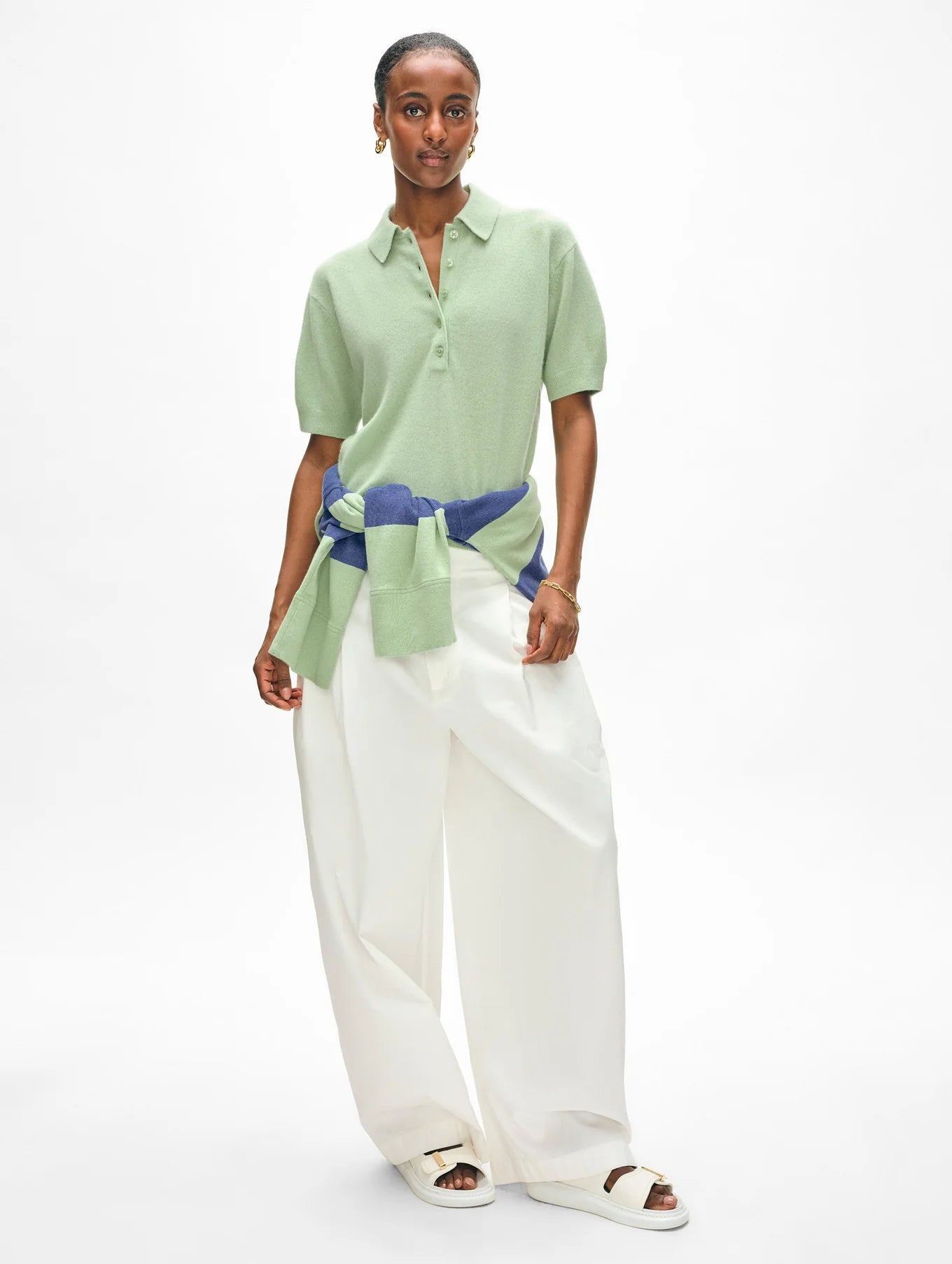 A woman wears a light green cashmere button polo by White + Warren with relaxed-fit white wide-leg pants. Her look is completed with white sandals and a retro-inspired blue and green sweater tied around her waist against a plain white background.