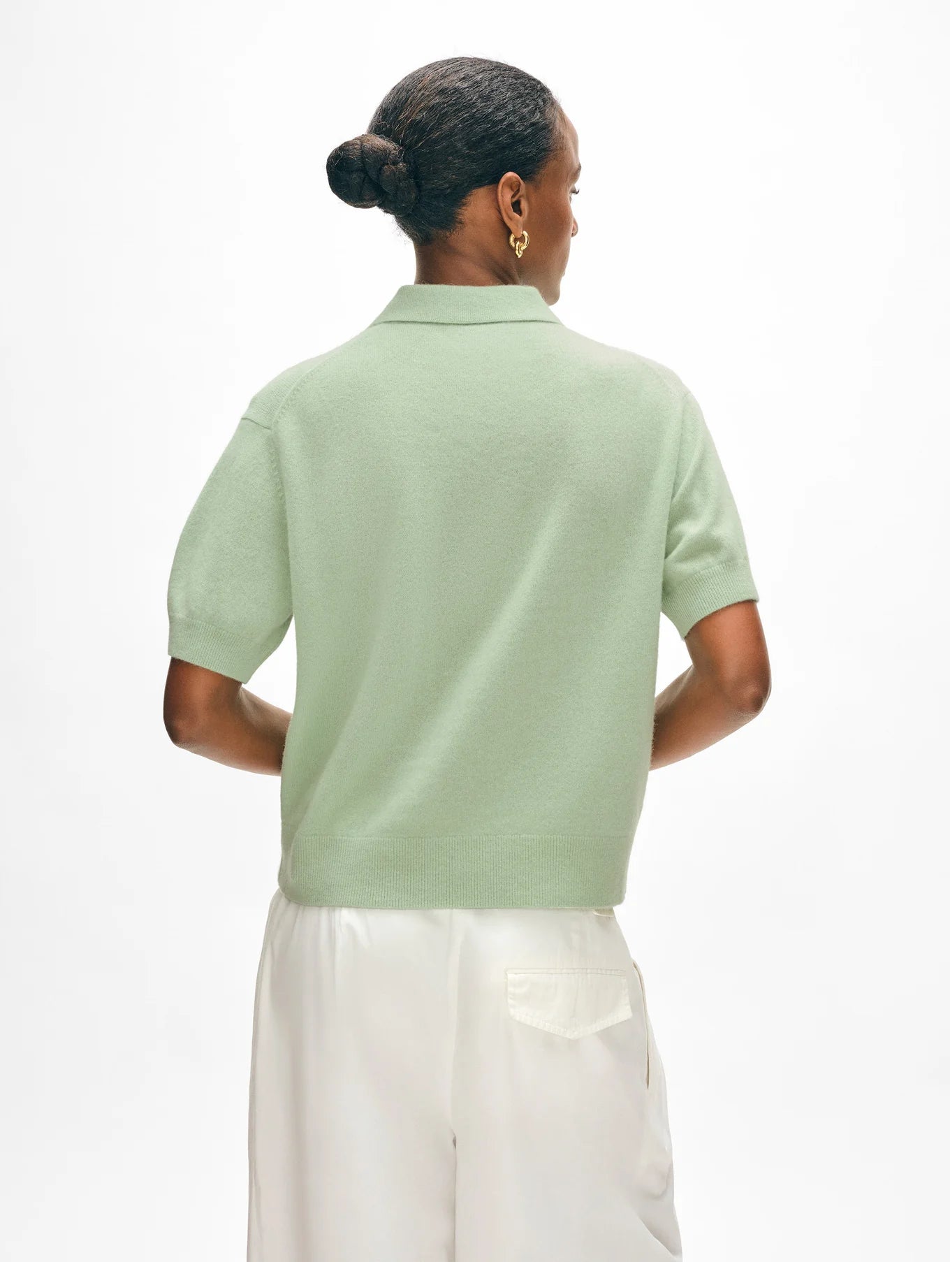 A person with short hair styled in a bun wears a light green cashmere button polo by White + Warren paired with white pants, facing away. The relaxed fit complements their gold hoop earrings against the plain white background.