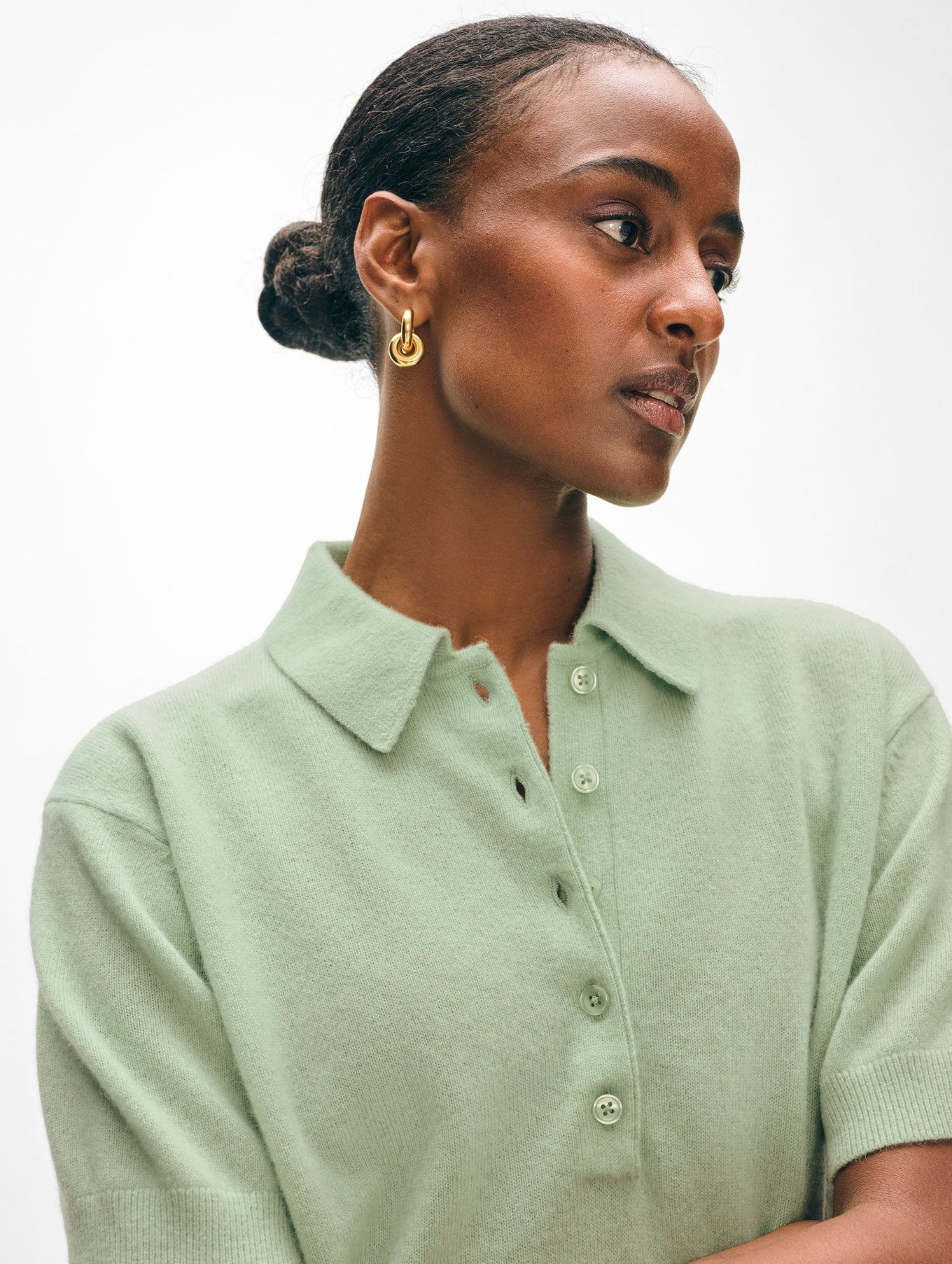 A person in a relaxed fit light green cashmere button polo by White + Warren gazes sideways, with their hair in a neat bun and gold hoop earrings. The background is soft white.