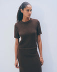 A woman with curly hair tied back wears the White + Warren Luxe Cotton T-shirt in dark brown, paired with a matching skirt, standing against a plain light blue wall and gazing to the side with a neutral expression.
