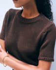 A woman with curly hair, wearing the Luxe Cotton T-shirt by White + Warren and a gold chain bracelet, stands with her arms crossed against a light background. Her face is partially visible.