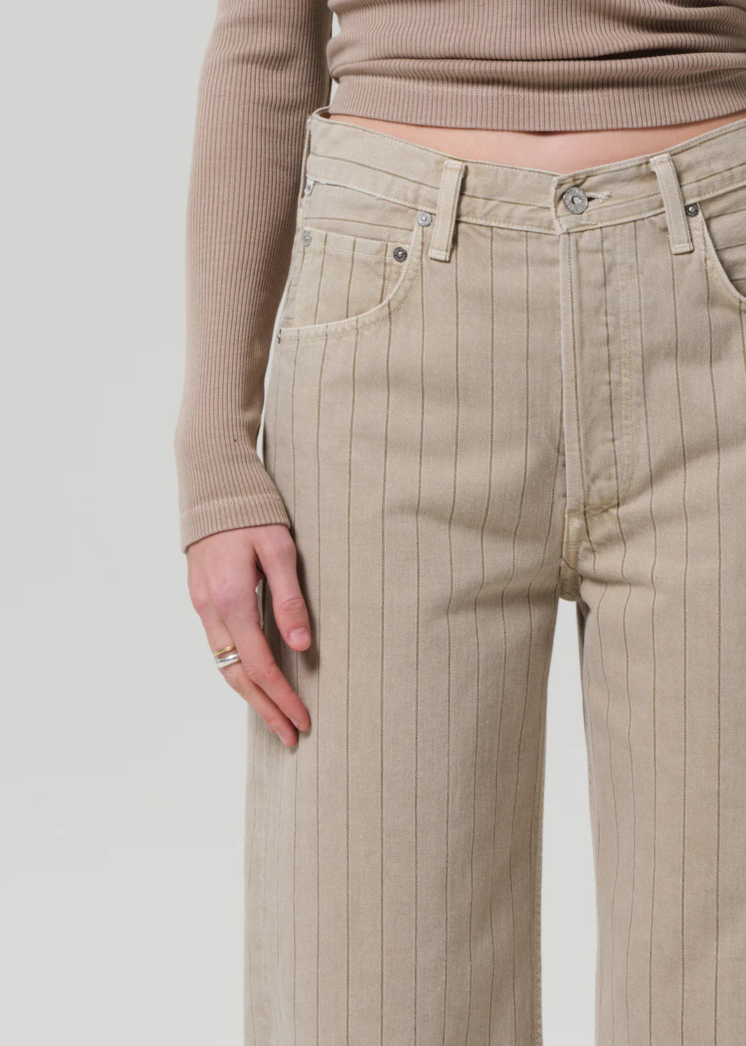 A person wears the Ayla Raw Hem Crop by Citizens Of Humanity/AGOLDE: beige, high-waisted, wide-leg pinstripe pants in heavyweight non-stretch cotton. Only their lower torso and one hand are shown against a plain light background.