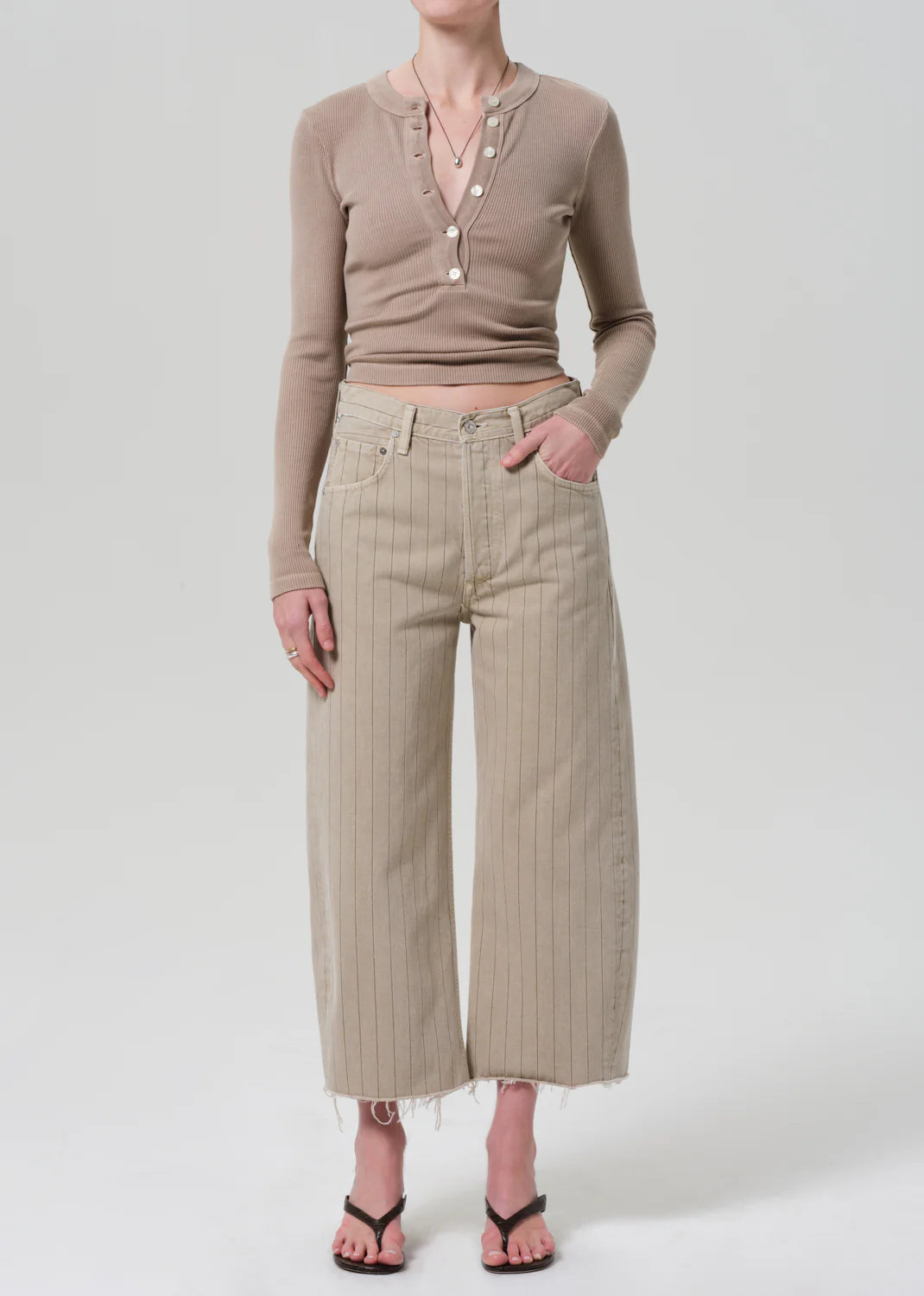 A person wears a fitted beige buttoned long-sleeve top, Citizens Of Humanity/AGOLDE Ayla Raw Hem Crop high-waisted wide-leg beige pinstripe pants in heavyweight cotton, and black heeled sandals, standing against a plain light background.