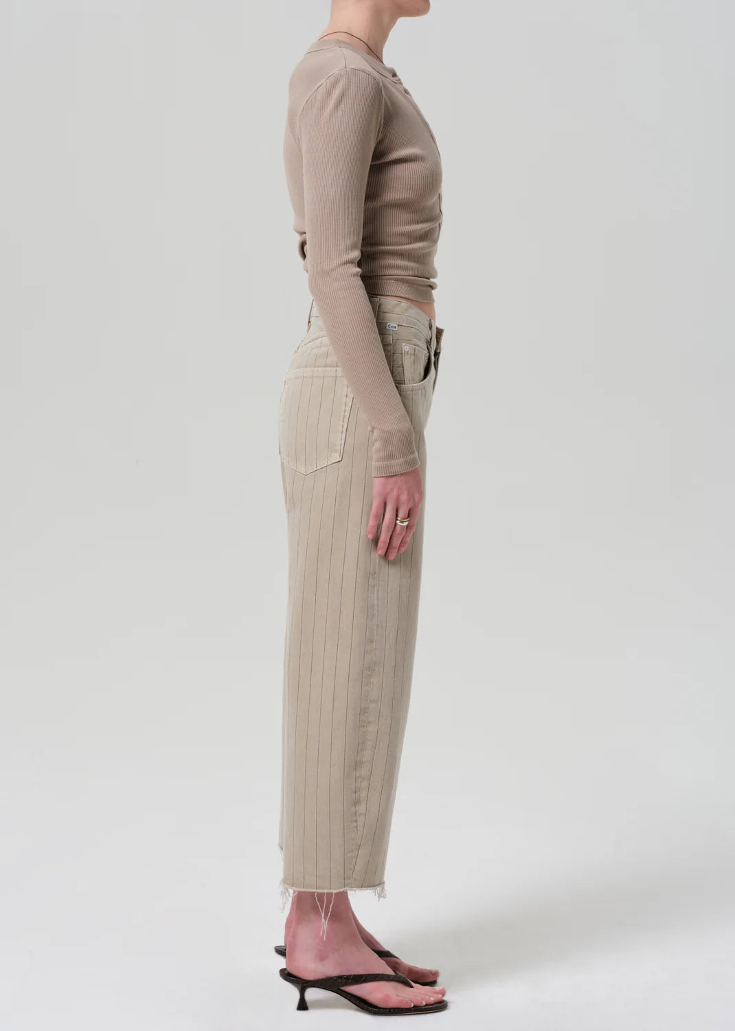 A person stands in profile against a light background, wearing a fitted beige long-sleeve top and high-waisted Citizens Of Humanity/AGOLDE Ayla Raw Hem Crop pinstripe pants in heavyweight non-stretch cotton with black heeled sandals.